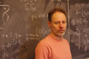 A man stands in front of a chalkboard. There are mathematical equations on the chalkboard.