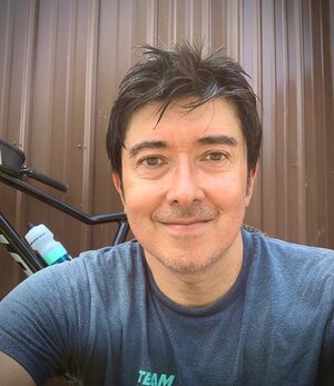 A man in a t-shirt poses in front of a bicycle and a building with metal siding. 